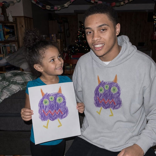 Big brother in a sport grey hoodie printed with a kid's monster drawing, next to the paper original