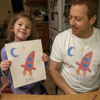 Dad in a white tee printed with a kid's rocket drawing, next to the paper original