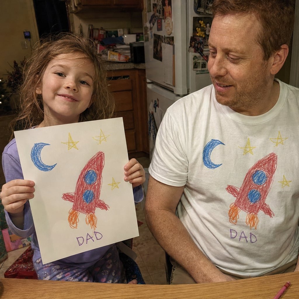 Dad in a white tee printed with a kid's rocket drawing, next to the paper original