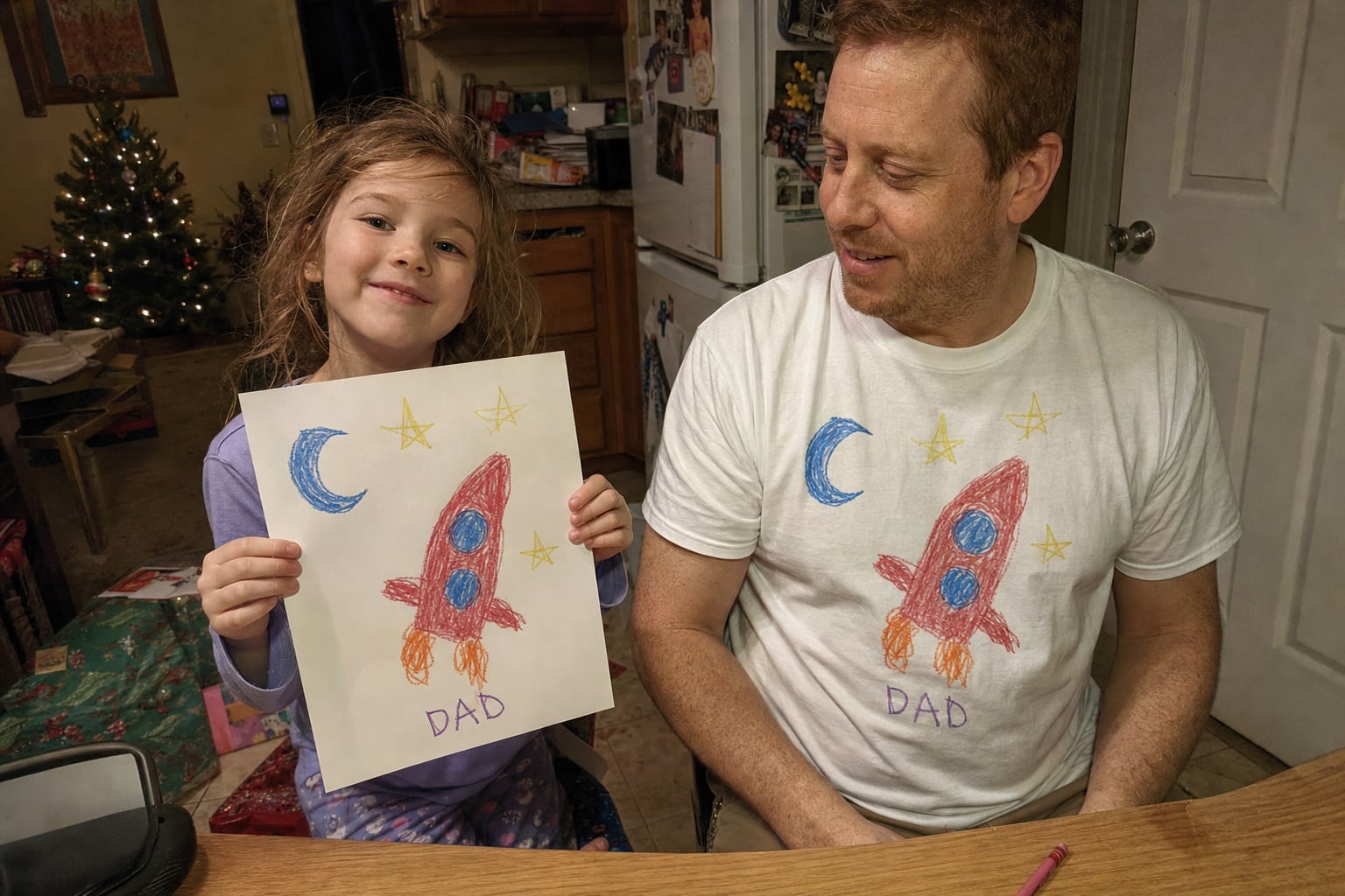 Dad in a white tee printed with a kid's rocket drawing, next to the paper original