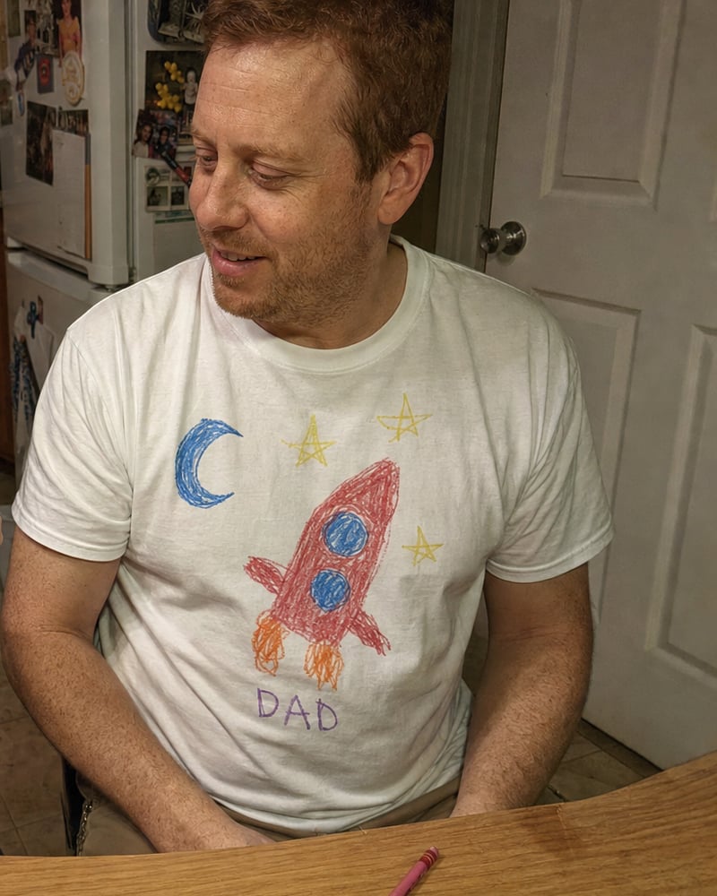 Dad in a white tee printed with a kid's rocket drawing, next to the paper original
