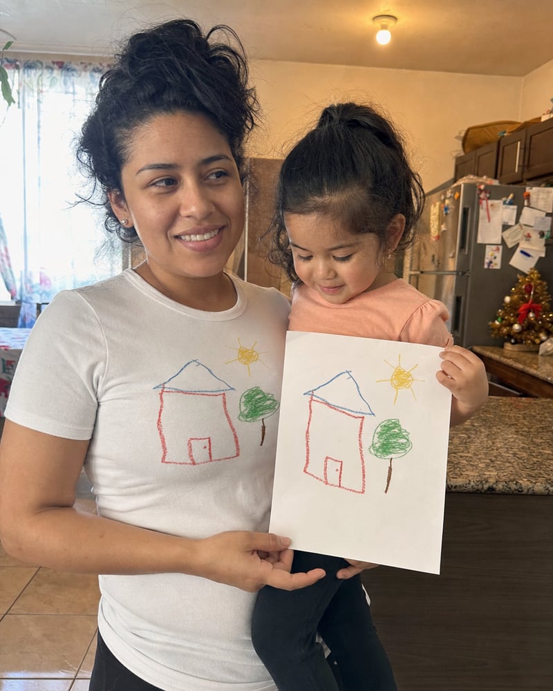Mom in a white tee printed with a kid's house drawing, next to the paper original
