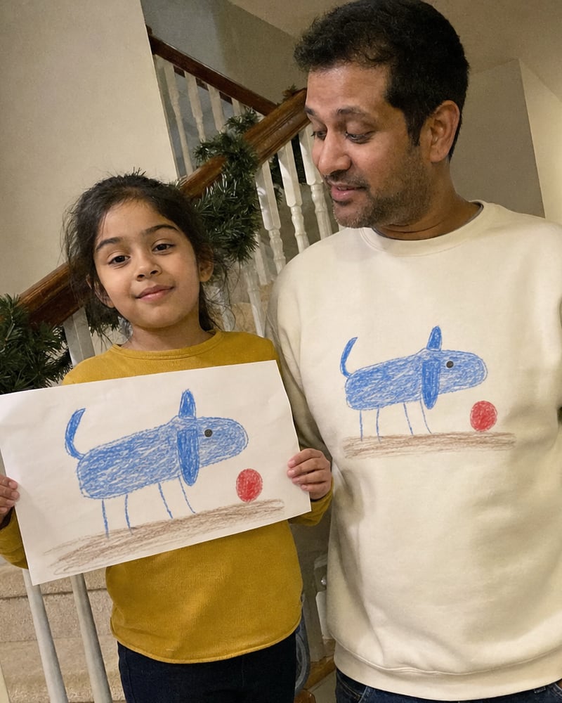 Dad in a sand sweatshirt printed with a kid's blue dog drawing, next to the paper original