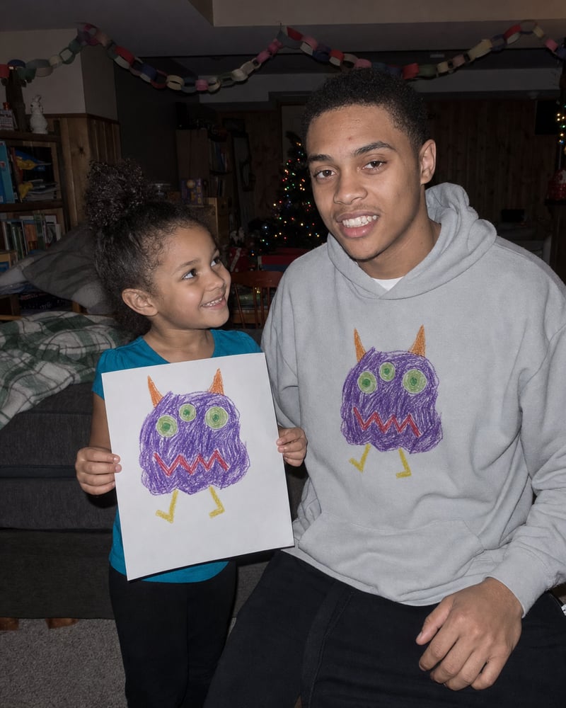 Big brother in a sport grey hoodie printed with a kid's monster drawing, next to the paper original